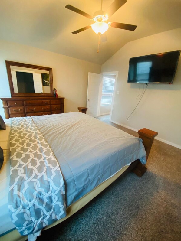 A bedroom with a bed, dresser, TV, ceiling fan, and a door leading to another room.