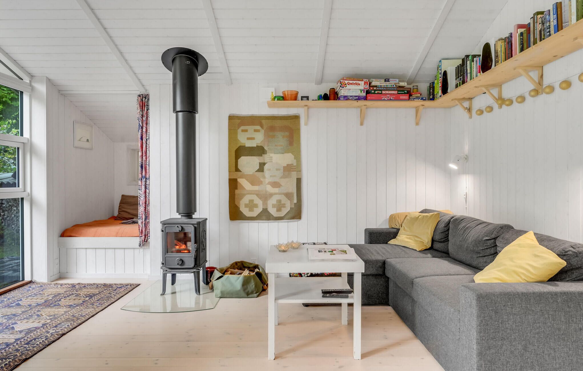 En hyggelig stue med en brændeovn, en grå sofa med gule puder, et hvidt sofabord og en reol fyldt med bøger og spil.