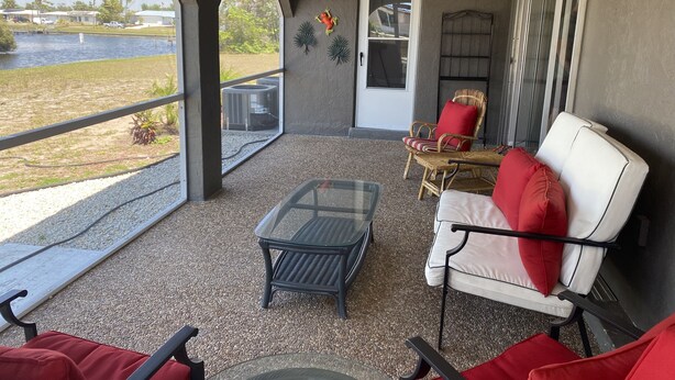 Uma área de estar coberta ao ar livre, com mesa de vidro, cadeiras de vime e vista para a água e vegetação.