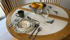 Uma mesa de madeira redonda com um prato verde e branco, uma caneca verde, um copo de água, uma tigela de fruta e talheres.