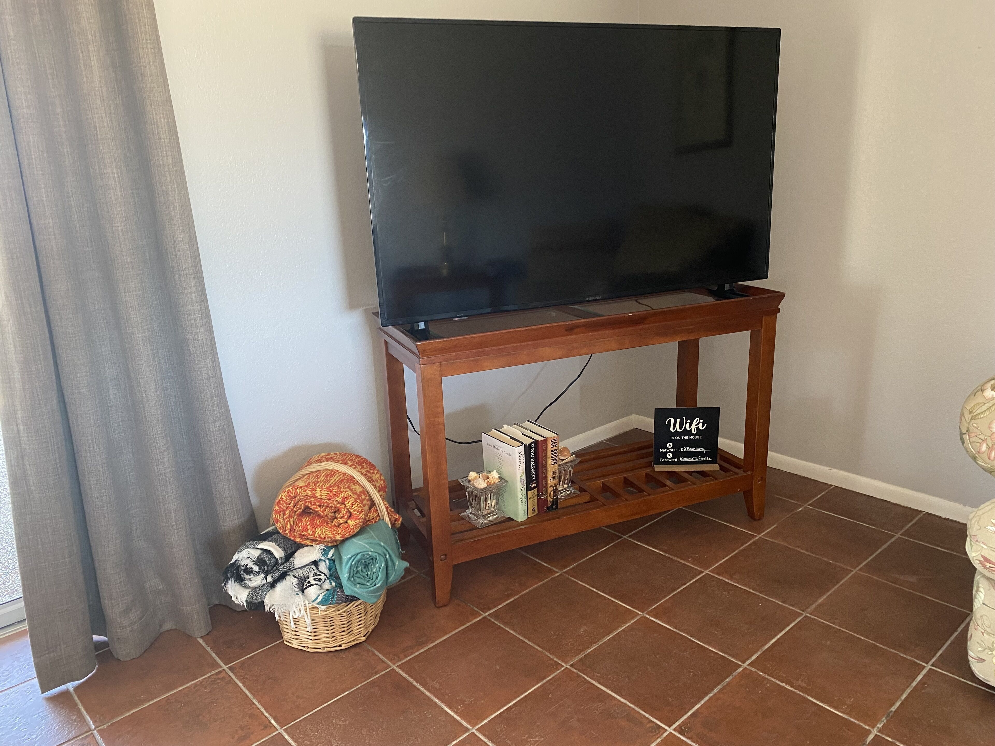 Sala de estar con televisor sobre un mueble de madera, una cesta con mantas y un cartel que indica la disponibilidad de WiFi.