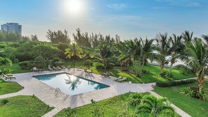 A swimming pool surrounded by lush greenery and palm trees, with lounge chairs and a clear sky. - Beautiful Condo on the Beach in Freeport! (Freeport)