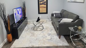 Uma sala de estar moderna com um sofá cinzento, uma mesa de centro de vidro e uma TV de tela plana sobre um suporte preto.