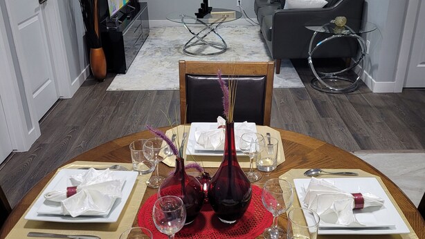Uma sala de jantar com uma mesa redonda posta para quatro pessoas, com um centro de mesa vermelho, guardanapos brancos e talheres. Uma mesa de centro de vidro com design moderno está ao fundo, e um sofá cinza é visível na sala de estar adjacente.