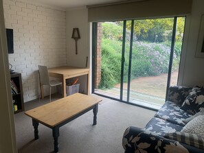Relax in this serene space with a sofa, wooden table, and bookshelf, leading out to a garden through a large sliding glass door.