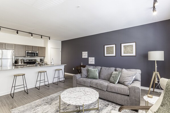 A modern living room with a grey sofa, a round coffee table, and a kitchen area with a microwave and bar stools.