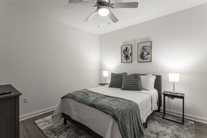 A bedroom with a bed, two bedside tables with lamps, a ceiling fan, and two wall-mounted artworks.