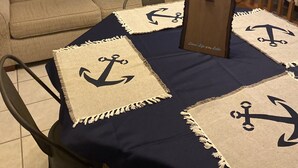 Un comedor con una mesa dispuesta para cuatro, con mantel azul marino y cuatro individuales con diseños de anclas. Se ve una silla de madera y una ventana que ofrece vistas al exterior.