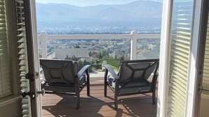 Una porta vetrata scorrevole aperta che dà su un balcone con due sedie e un tavolo. Il balcone si affaccia su una zona residenziale con case e montagne sullo sfondo.