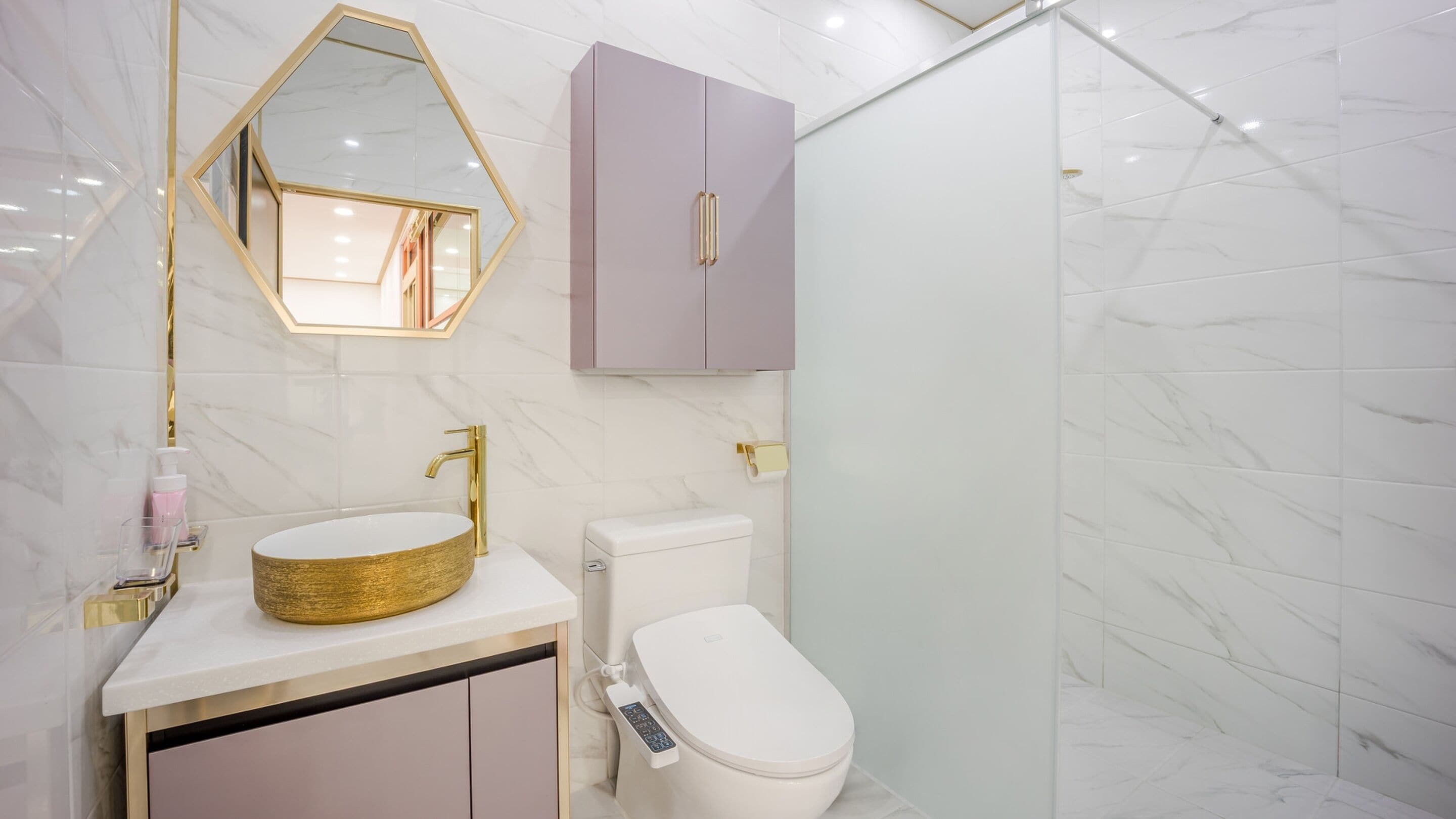 Une salle de bain moderne avec un sol en marbre, un lavabo rond doré, des toilettes dotées d’un panneau de commande et un meuble violet.