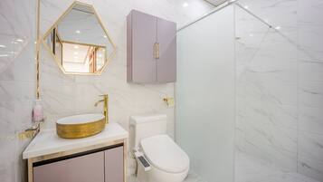Une salle de bain moderne avec un sol en marbre, un lavabo rond doré, des toilettes dotées d’un panneau de commande et un meuble violet.