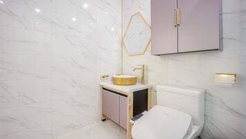 Un baño moderno con suelo de mármol, un lavabo redondo con grifo dorado, un inodoro y un mueble con espejo.