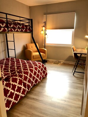 A dormitory room with a bunk bed, a desk, a chair, and a window with blinds.
