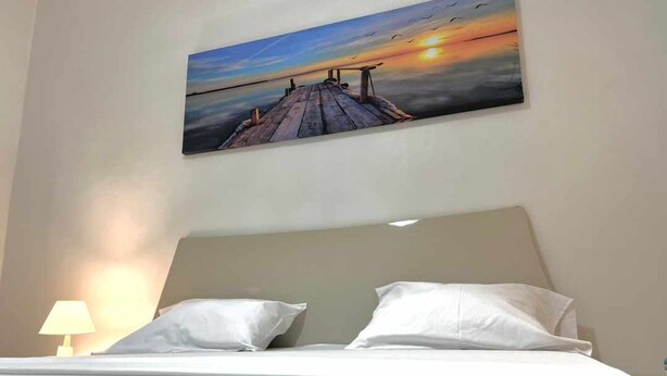 A bedroom with a bed, two pillows, a nightstand with a lamp, and a painting of a sunset over a pier. - Apartement Meublé à Loué aux Almadies (Dakar)