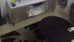 A living room with a flat-screen TV on a wooden cabinet, a small plant on a shelf, and a black round table. - Apartement Meublé à Loué aux Almadies (Dakar)