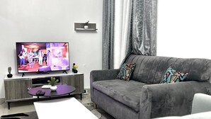 A living room with a grey sofa, a flat-screen TV, a small table with a potted plant, and a coffee table. - Apartement Meublé à Loué aux Almadies (Dakar)