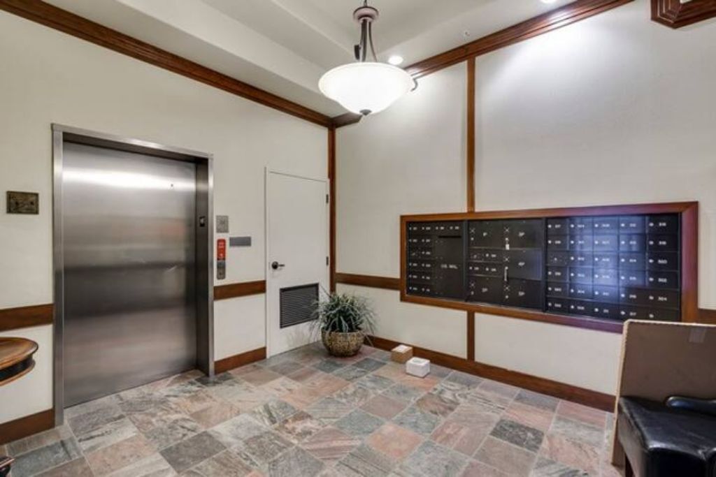 A lobby with a metallic elevator, a bulletin board, a potted plant, and a seating area.
