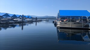 A marina with boats docked under blue canopies, calm water, and a mountainous backdrop.
