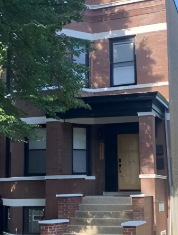 Un edificio de ladrillo de dos pisos con puerta principal, ventanas y un pequeño porche.
