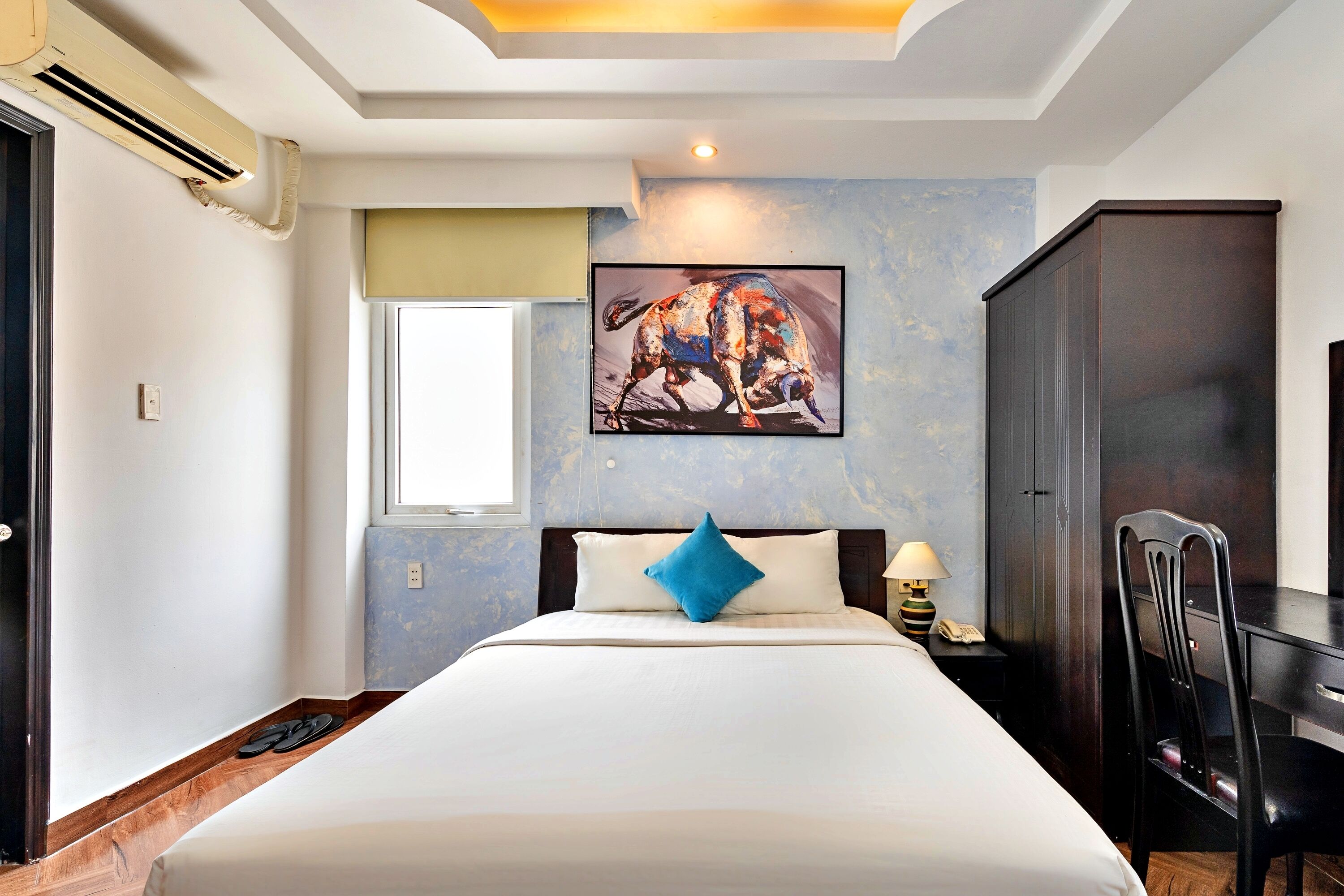 A hotel room with a large bed, a desk, a chair, a painting on the wall, and an air conditioning unit.