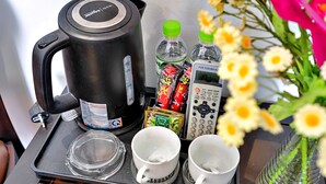 A tray with a black electric kettle, two empty cups, a glass, a bottle of water, a pack of tea, a pack of snacks, a remote control, and a vase with flowers. - S79 Residences Yaya Bui Vien (Ho Chi Minh City)