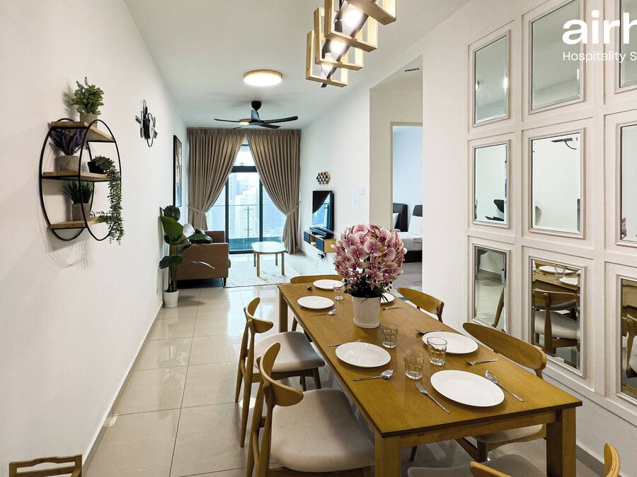 A hotel dining area with a long wooden table set for four, surrounded by chairs. There is a ceiling fan above the table, and a potted plant on the left side.
