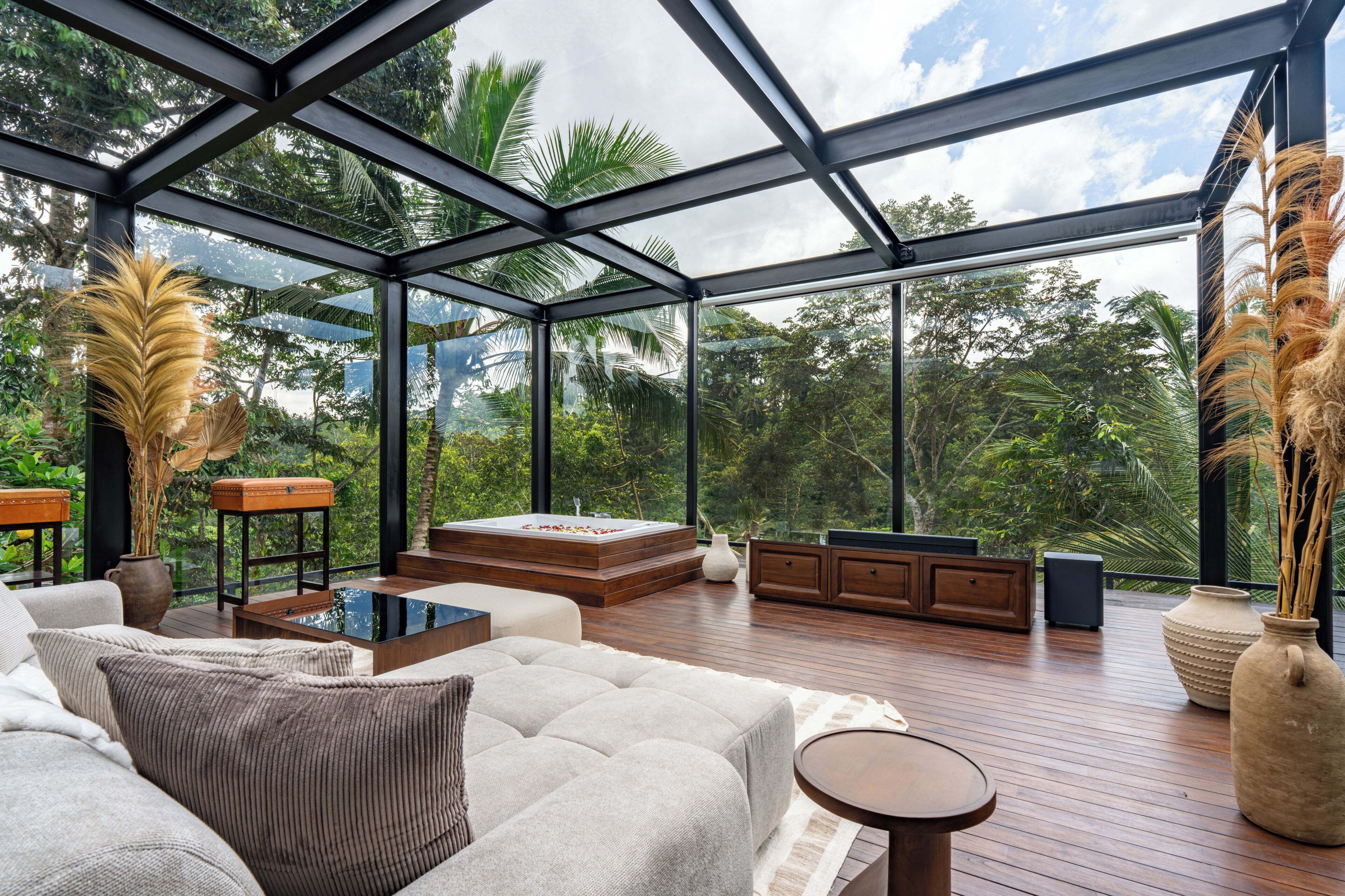 A spacious room with a glass ceiling, a large sofa, a wooden deck, and a view of lush greenery.