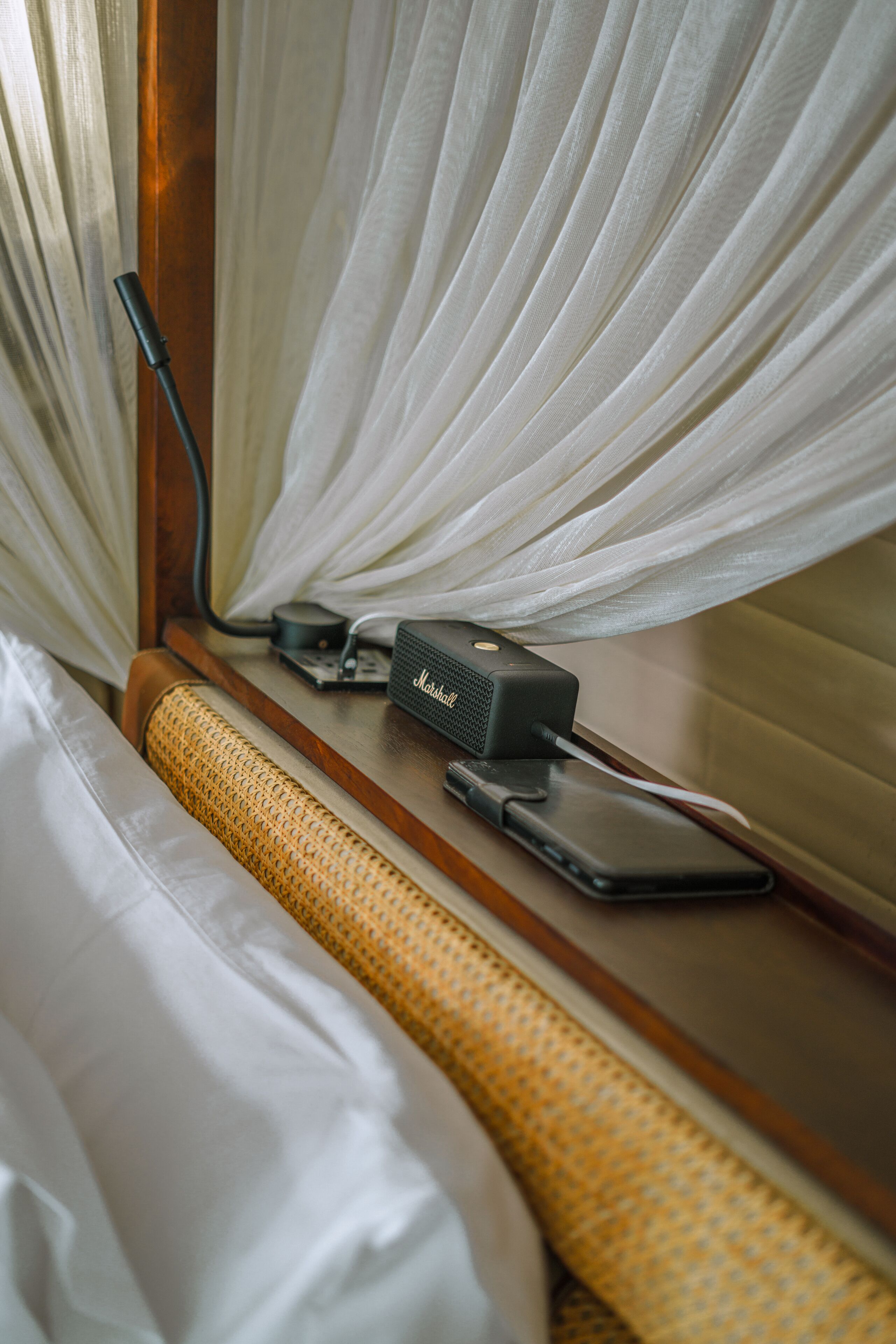 A bed with white linens, a woven headboard, a bedside table with a power bank, a phone, and a speaker.