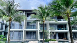 Un immeuble moderne à plusieurs étages, doté de balcons, d’un espace piscine et de palmiers.