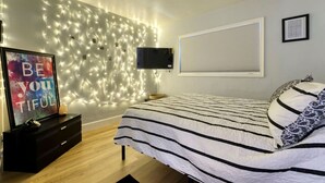 Une chambre à coucher avec un lit, un téléviseur fixé au mur, des guirlandes lumineuses, une affiche motivante et une fenêtre avec des stores.