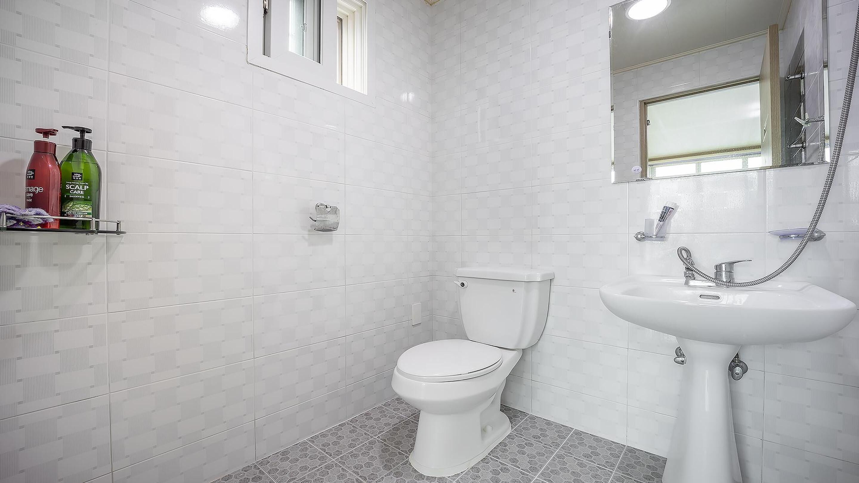 Un baño con paredes de azulejos blancos, inodoro blanco, lavamanos con ducha de mano y una ventana sobre el inodoro.