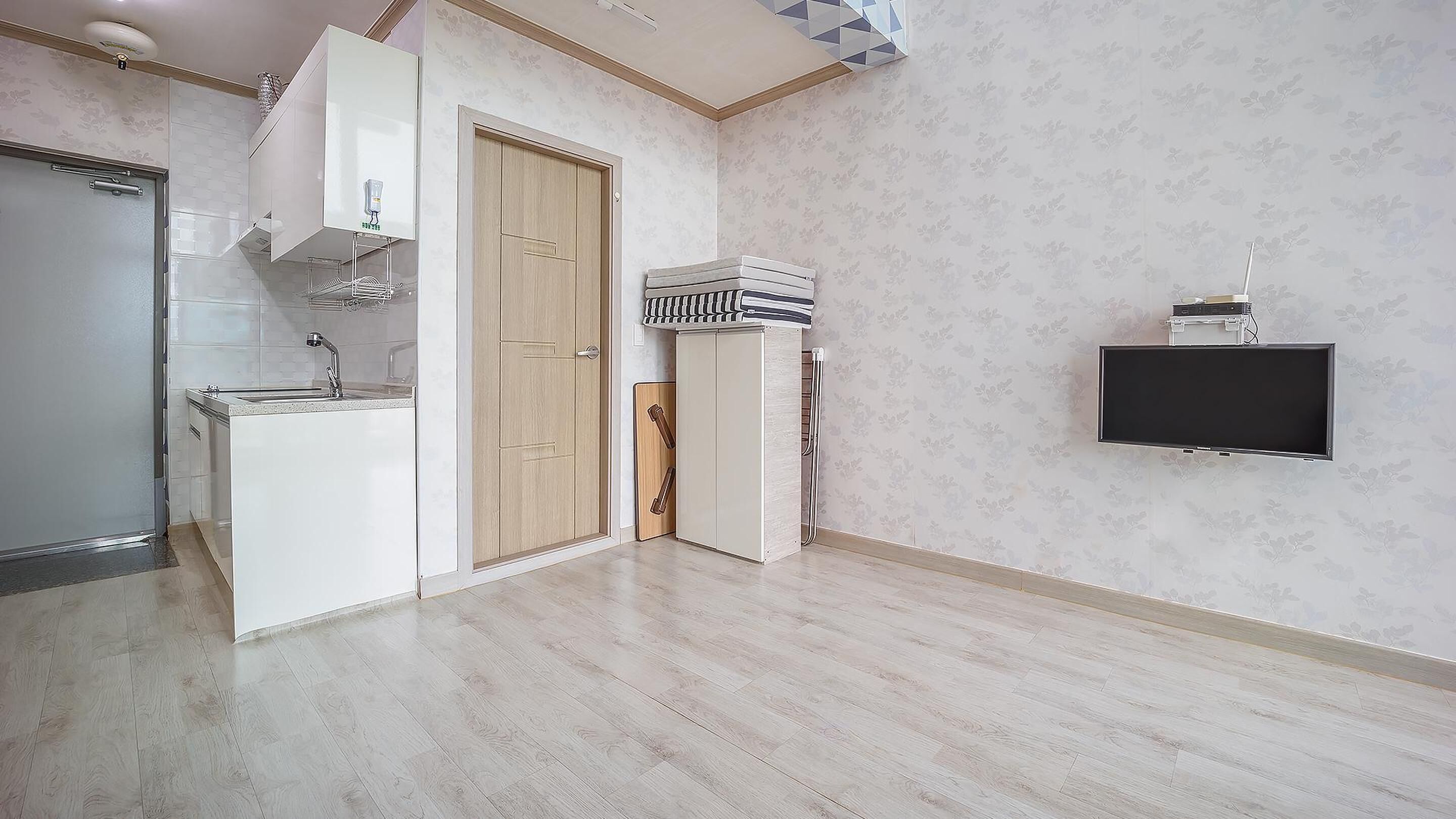 Una cocina moderna con horno empotrado, microondas y lavamanos. Se observa una puerta de madera y un mueble pequeño con una toalla a rayas. Un televisor de pantalla plana está montado en la pared.
