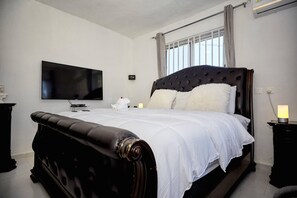 A bedroom with a large bed, a flat-screen TV mounted on the wall, a bedside table with a lamp, and a window with curtains.