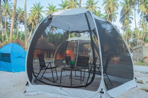 Una tienda en forma de cúpula con techo transparente, rodeada de sillas y una pequeña mesa, situada en una playa arenosa con palmeras al fondo.