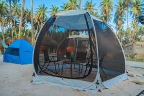 A dome tent with a table and chairs inside, set up on a sandy beach with palm trees in the background.