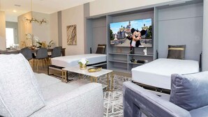 A modern hotel room with two beds, a dining area, and a TV displaying a cartoon character.
