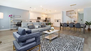 A modern living room with a grey sofa, a dining area with a table and chairs, and a kitchen in the background.