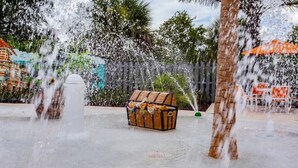 A water fountain with a treasure chest in the center, surrounded by palm trees and a white fence.