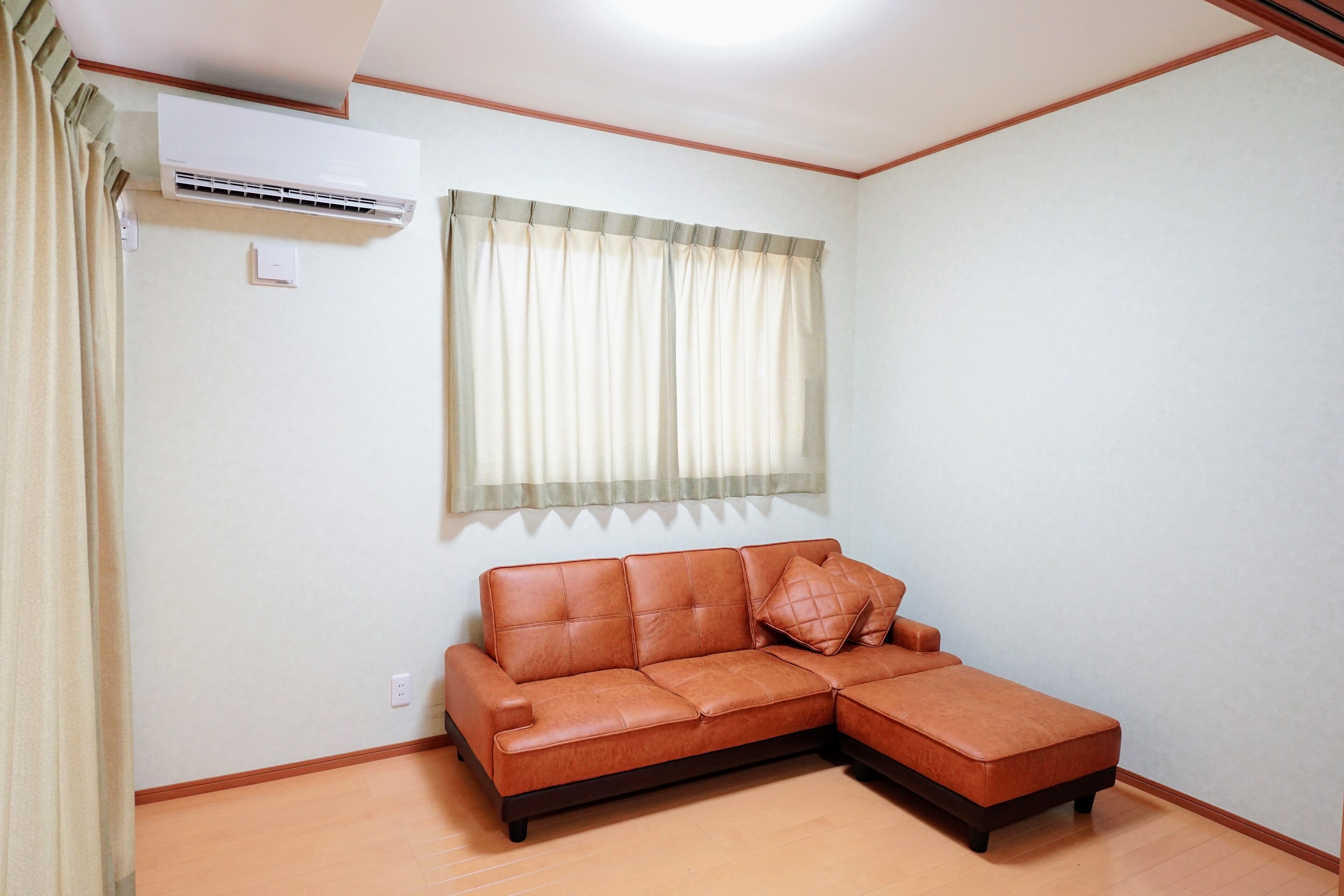 Ein Zimmer mit einem braunen Ledersofa, einem Fenster mit durchscheinenden Vorhängen und einer an der Wand befestigten Klimaanlage.