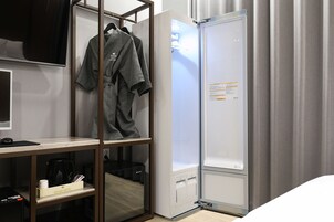 A hotel room with a bed, a desk with a computer, a coffee machine, and a wardrobe with a robe hanging.
