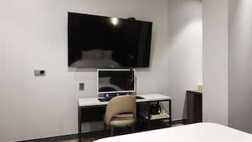 A hotel room with a bed, a desk with a computer, a chair, and a TV mounted on the wall.