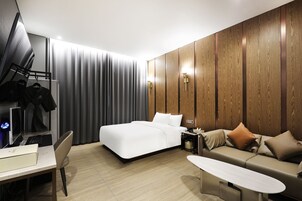 A modern hotel room with a bed, a sofa, a desk with a computer, and a wardrobe.