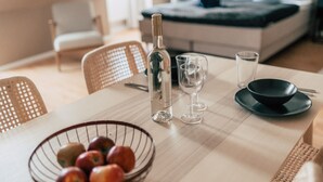 A dining table with a bottle, two glasses, a bowl of fruit, and a black bowl on a light-colored tablecloth. - Luga Homes - Rosental (Leipzig)