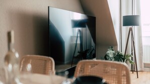 A dining area with a table, chairs, a television, a floor lamp, and a potted plant. - Luga Homes - Rosental (Leipzig)