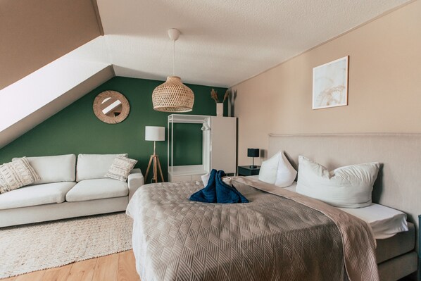 A modern bedroom with a sloped ceiling, a bed with a patterned bedspread, a white sofa with cushions, a standing lamp, and a framed picture on the wall.