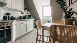 A kitchen with white cabinets, a stove, and a dining area with wicker chairs and a table. - Luga Homes - Rosental (Leipzig)