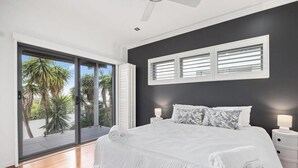 A bedroom with a large bed, two bedside tables, a window with blinds, and a view of palm trees. - Sandy View - 180 degree views - Luxury Home (Nikenbah)