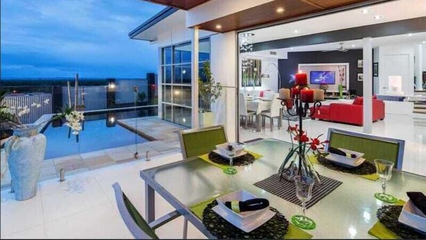A modern outdoor dining area with a glass table, chairs, and a view of the cityscape at dusk. - Sandy View - 180 degree views - Luxury Home (Nikenbah)