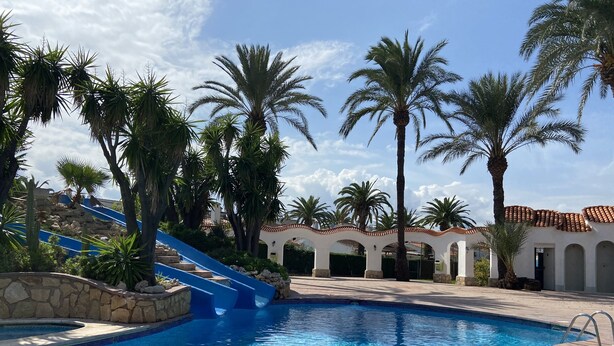 A swimming pool with a blue slide, surrounded by palm trees and a building with arches. - Beautiful oasis with pool landscape near the dream beach Las Marinas (Dénia)