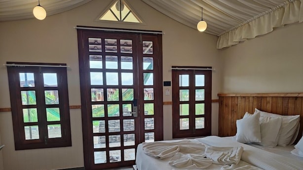 A bedroom with a large bed, wooden headboard, and two windows with views of greenery. - Nirvana Resort and Restaurant (Garudeshwar)